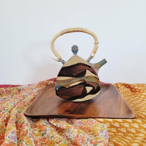 Puede incluir: Tetera de cerámica con asa de bambú, con un diseño abstracto en tonos marrón, beige y azul. La tetera se encuentra sobre una bandeja de madera. Una pieza decorativa para los amantes del té. Altura aproximada: 20 cm.