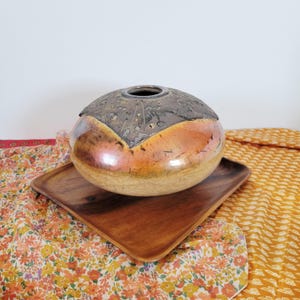 May include: A decorative ceramic vase with a metallic, patterned lid. The vase has a rounded shape with a warm, iridescent glaze in shades of orange and brown. It sits on a square wooden tray, showcasing a unique home decor item.