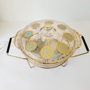 Puede incluir: Una fuente para horno vintage de vidrio transparente con tapa, con detalles dorados y turquesas. La fuente tiene un soporte de metal dorado y asas negras. La tapa está adornada con diseños circulares turquesas y dorados.