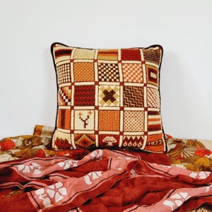 Könnte beinhalten: Quadratisches Zierkissen mit einem Patchwork-Design in Braun-, Orange- und Cremetönen. Das Kissen hat einen schwarzen Rand und liegt auf einem Stoff mit Blumenmuster. Das Kissen zeigt verschiedene geometrische Muster.