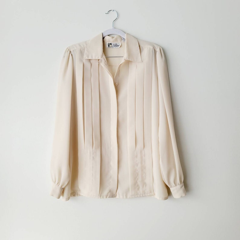 Op de afbeelding: Cr&egrave;mekleurige blouse met lange mouwen, een kraag en een geplooide voorkant. De blouse heeft een knoopsluiting en hangt aan een zilveren hanger. De mouwen hebben manchetten met knopen. De blouse is gemaakt van een lichte stof.