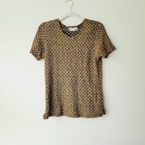 May include: A black and gold short-sleeved shirt with a floral pattern. The shirt has a round neckline and is hanging on a white hanger.