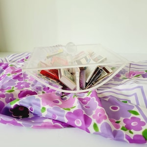 Puede incluir: Una caja de acrílico transparente con tapa, con forma de semicírculo, llena de diferentes cajas de cerillas. La caja está sobre una tela floral de color morado y blanco.