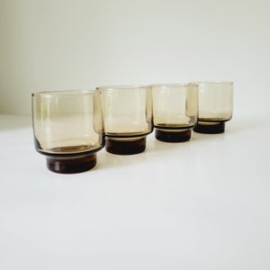 May include: Four vintage amber-colored drinking glasses. Each glass has a cylindrical shape with a dark brown base. The glasses are arranged in a row, showcasing their simple, yet elegant design. Perfect for serving beverages.