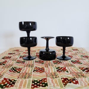 Puede incluir: Disposición de cuatro copas de tallo negro, incluidas dos apiladas, sobre un mantel estampado. Las copas tienen forma de cuenco redondeado y un tallo delgado. El mantel presenta un diseño geométrico repetitivo en rojo, verde y beige.