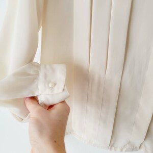 Op de afbeelding: Cr&egrave;mekleurige blouse met lange mouwen en een manchet met knoop. De blouse heeft verticale plooien aan de voorkant en een klassiek, elegant ontwerp. De manchet heeft een enkele, kleine parelknop.