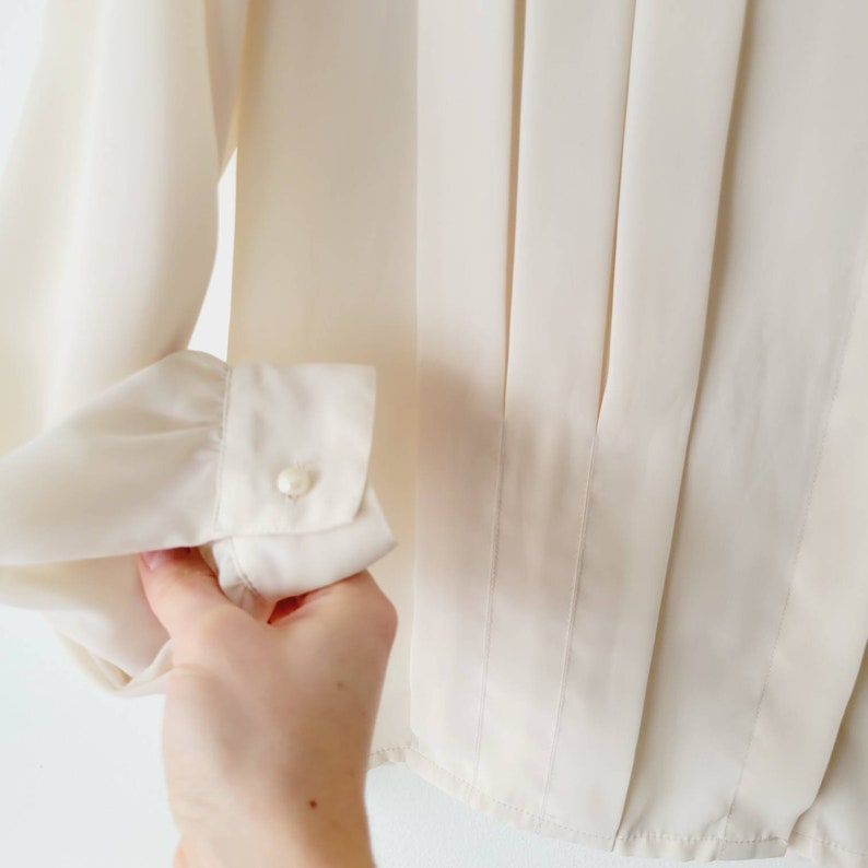 Op de afbeelding: Cr&egrave;mekleurige blouse met lange mouwen en een manchet met knoop. De blouse heeft verticale plooien aan de voorkant en een klassiek, elegant ontwerp. De manchet heeft een enkele, kleine parelknop.