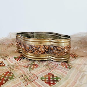 May include: A decorative brass planter with a wavy rim and a copper-colored floral design. The planter is sitting on a patterned fabric.