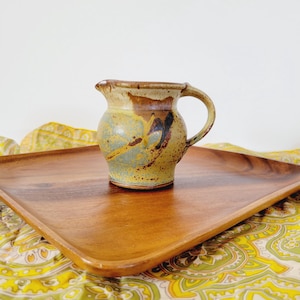 May include: A small, handcrafted ceramic pitcher with a handle. The pitcher features a speckled, earthy color palette of brown, tan, and blue-green, with dark brushstrokes. It sits on a wooden tray.