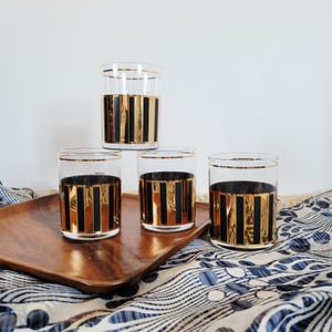 May include: Four clear drinking glasses with gold and black vertical stripes. The glasses are arranged on a wooden tray with a blue and white patterned cloth in the background. Each glass has a gold rim.