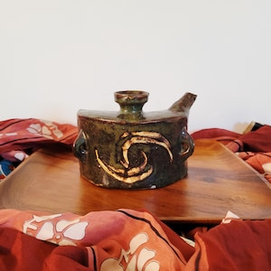 May include: A handmade ceramic teapot with a dark green glaze and a swirling, abstract design in cream. The teapot has a spout, a handle, and a small, decorative top. It sits on a wooden tray.