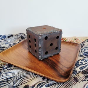 Puede incluir: Un dado grande de metal hecho a mano con agujeros circulares perforados, colocado sobre una bandeja de madera. El dado es de color marrón oscuro y desgastado con marcas de soldadura visibles. La bandeja es de color marrón medio.