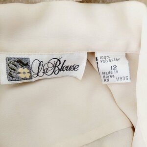 Op de afbeelding: Cr&egrave;mekleurige blouse met een label met de tekst "La Blouse". Het label geeft aan dat de blouse 100% polyester is, maat 12 en gemaakt in Korea. De blouse heeft een zachte, vloeiende textuur.