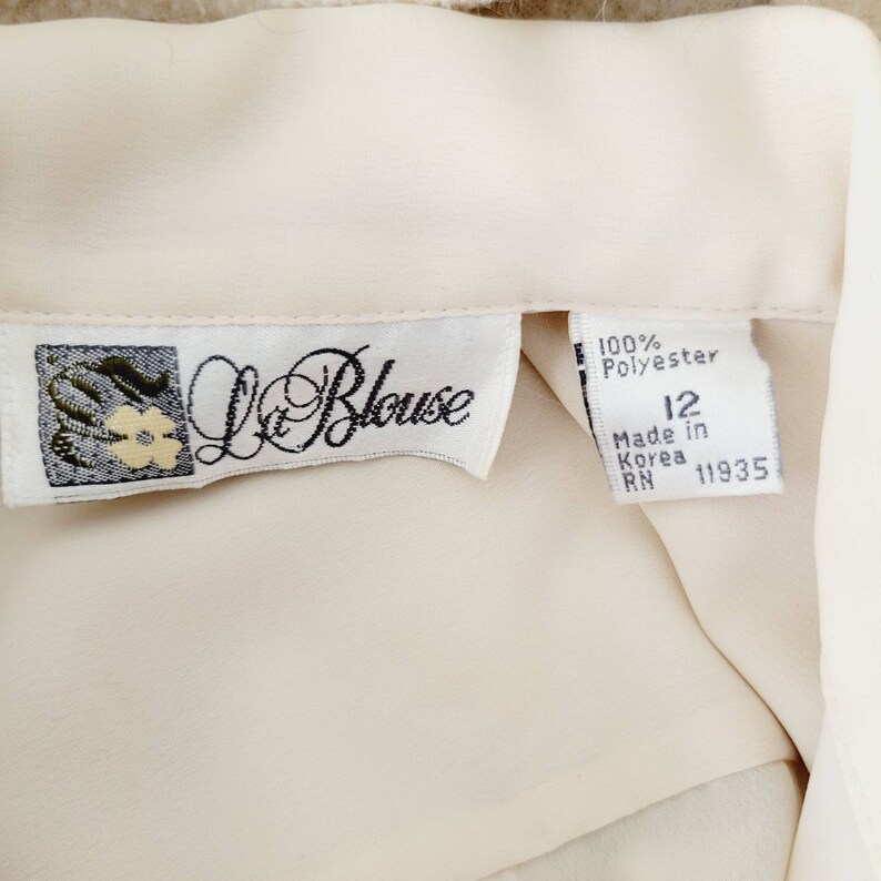 Op de afbeelding: Cr&egrave;mekleurige blouse met een label met de tekst "La Blouse". Het label geeft aan dat de blouse 100% polyester is, maat 12 en gemaakt in Korea. De blouse heeft een zachte, vloeiende textuur.