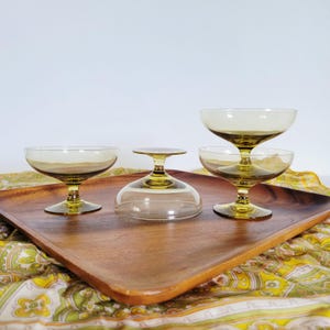 Könnte beinhalten: Vier Vintage-Champagner-Coupe-Gläser mit bernsteinfarbenen Schalen und Stielen, präsentiert auf einem Holztablett. Ein Glas ist umgedreht. Die Gläser haben ein klassisches, elegantes Design, ideal zum Servieren von Cocktails oder Desserts.