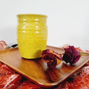 Pode incluir: Um vaso de cerâmica amarela com acabamento craquelado está sobre uma bandeja de madeira. Duas rosas vermelhas secas estão ao lado do vaso. A bandeja repousa sobre um tecido estampado em vermelho e branco, criando uma exibição rústica e artística.