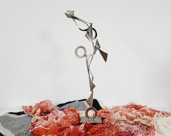 Könnte beinhalten: Eine silberne Metallskulptur mit geometrischen Formen, die auf einem Sockel steht. Die Skulptur ist auf einem einzigen Punkt balanciert und scheint sich zu bewegen. Sie wird auf einem roten und weißen gemusterten Stoff präsentiert.