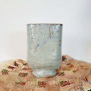 May include: A light blue ceramic vase with a textured surface and a wide, cylindrical shape. The vase has a slightly flared base and a rough, uneven texture with streaks of blue and brown. The background is white.