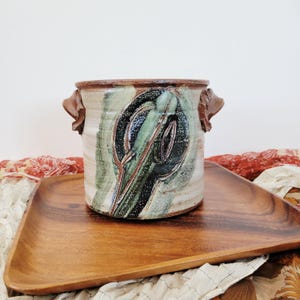May include: A cylindrical ceramic crock with two side handles, featuring a cream-colored base glaze with a prominent dark green and black abstract brushstroke design. The rim and handles are finished in a contrasting reddish-brown clay tone.