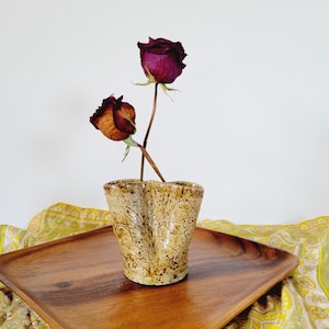 Puede incluir: Un pequeño jarrón de cerámica beige con una superficie texturizada y dos aberturas, que contiene dos rosas secas. Las rosas son de color burdeos intenso y naranja. El jarrón está sobre una bandeja de madera.