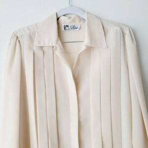 Op de afbeelding: Cr&egrave;mekleurige blouse met knopen en verticale plooien. De blouse heeft een kraag en lange mouwen. Een klein label is zichtbaar bij de halslijn. De blouse hangt aan een witte hanger tegen een witte achtergrond. De blouse is gemaakt van een lichte stof.