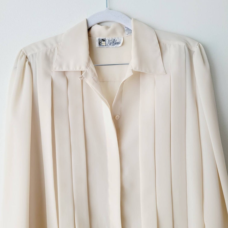 Op de afbeelding: Cr&egrave;mekleurige blouse met knopen en verticale plooien. De blouse heeft een kraag en lange mouwen. Een klein label is zichtbaar bij de halslijn. De blouse hangt aan een witte hanger tegen een witte achtergrond. De blouse is gemaakt van een lichte stof.