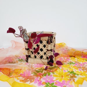 May include: A square-shaped macrame plant pot with a brown wooden frame and a natural rope design. The pot is filled with a trailing purple plant and is sitting on a colorful floral fabric.