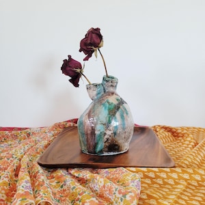Puede incluir: Un jarrón de cerámica hecho a mano con una forma orgánica única. El jarrón presenta una mezcla de esmalte turquesa, marrón y blanco. Dos rosas rojas secas se colocan en el jarrón. Se encuentra sobre una bandeja de madera, con tela colorida en el fondo.