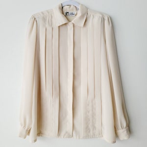 Op de afbeelding: Een cr&egrave;mekleurige blouse met verticale plooien aan de voorkant. De blouse met lange mouwen heeft een kraag en manchetten met knopen. De blouse hangt aan een witte hanger tegen een witte achtergrond. Het label luidt "J.B. Rose".