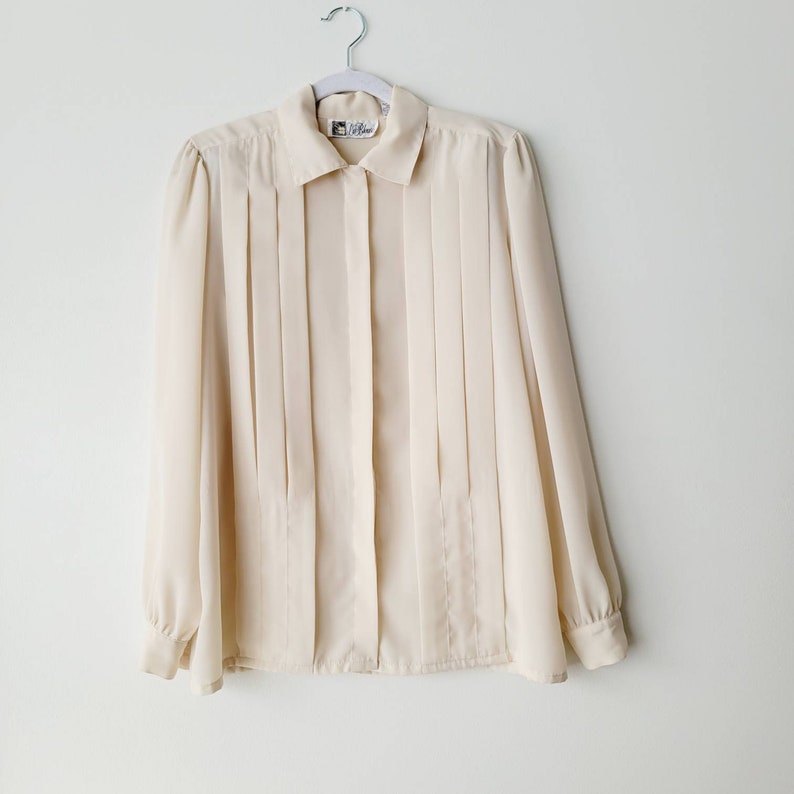 Op de afbeelding: Een cr&egrave;mekleurige blouse met lange mouwen, een kraag en een geplooide voorkant. De blouse hangt aan een witte hanger tegen een witte achtergrond. De mouwen hebben manchetten met knopen. Een klein label is zichtbaar bovenaan.