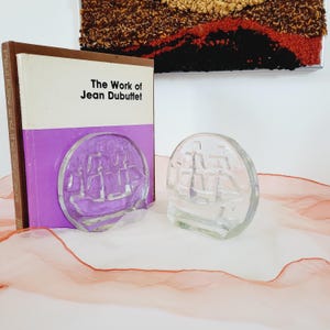 Puede incluir: Dos sujetalibros de vidrio con forma de barcos. Los sujetalibros son de vidrio transparente con un diseño esmerilado. Los sujetalibros están sobre una tela rosa y naranja. Los sujetalibros están al lado de un libro titulado "The Work of Jean Dubuffet".