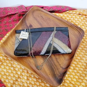 May include: A rectangular clutch bag featuring a geometric patchwork design of genuine snake skin in black, burgundy, and beige tones. The bag includes a thin gold-tone metal chain strap and a white tag with the text Genuine SNAKE attached.