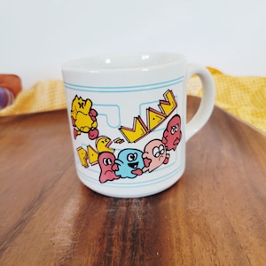 May include: White ceramic mug featuring a colorful Pac-Man design. The mug has a blue border and a handle. The Pac-Man characters are in yellow, pink, blue, and red. The mug has the words "PAC-MAN" in yellow.