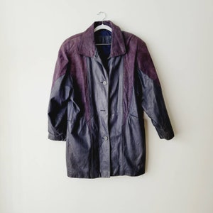 May include: A vintage leather coat in shades of purple and navy blue hangs on a white background. The coat features a collared neckline, button-down closure, and textured panels on the shoulders and sides. The coat is a mid-length style.