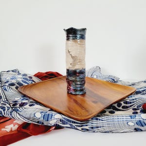 Könnte beinhalten: Eine handgefertigte Keramikvase mit strukturierter, schillernder Oberfläche. Die Vase hat einen dunklen, wellenförmigen Rand und einen Farbverlauf aus Blau, Lila und Rot. Sie steht auf einem quadratischen Holztablett, mit gemustertem Stoff im Hintergrund.