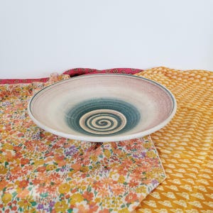 May include: A large, round ceramic bowl with a spiral design in the centre. The bowl features a gradient of colours, including white, pink, and teal. The bowl is placed on a floral patterned fabric.