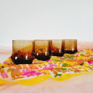 May include: Four vintage amber-colored drinking glasses with a unique, geometric base. The glasses are arranged on a colorful floral patterned scarf with pink, yellow, and orange hues. The background is a plain white.
