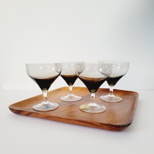 May include: Four vintage coupe glasses with smoky gray bowls and clear stems are arranged on a wooden tray. The glasses have a classic design, perfect for serving cocktails or champagne. The tray is made of a warm-toned wood.