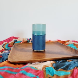 May include: A two-tone blue ceramic tumbler on a wooden tray. The top half of the tumbler is a light blue, with a darker blue base. The tray is placed on a colourful patterned fabric. The tumbler is likely handmade.