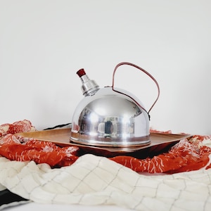 May include: A silver tea kettle with a red handle and a red spout sits on a wooden tray. The tray is on top of a red and white patterned fabric.