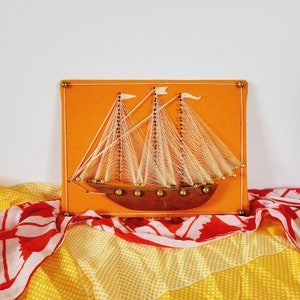 May include: A string art sailboat on an orange background. The sailboat is made of white string and has a brown wooden hull. The sailboat is surrounded by gold tacks.
