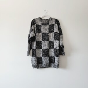 Op de afbeelding: Een zwart-wit gebreide trui met lange mouwen. De trui hangt aan een witte hanger tegen een witte achtergrond. De trui heeft een losse pasvorm en lijkt van gebreid materiaal te zijn.