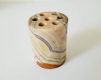 May include: A cylindrical toothbrush holder made of banded stone. The holder is beige, brown, and cream-colored with six holes on top. The stone has a natural, layered appearance. The holder is a decorative bathroom accessory.