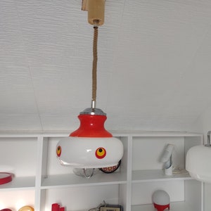 Puede incluir: Dos lámparas colgantes blancas y rojas con una fijación de techo blanca. Las lámparas colgantes tienen una pantalla blanca con detalles rojos y tres círculos rojos en la pantalla. Las lámparas cuelgan de un techo blanco.