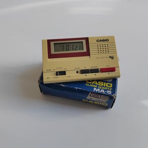 May include: A vintage Casio Alarm Quartz MA-5 digital clock, cream-coloured with a burgundy accent, displaying the time 10:21. The clock sits atop its original blue box, which has the Casio logo and product information.