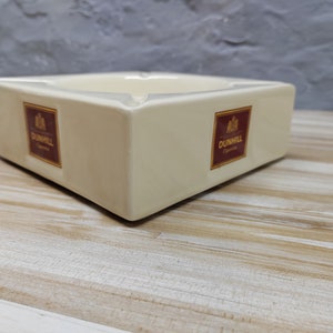 Op de afbeelding: Een witte asbak van keramiek met het Dunhill Cigarettes logo aan beide zijden. De asbak is rechthoekig met afgeronde hoeken en een kleine inkeping in het midden.
