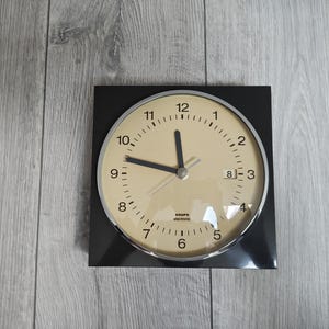 Può includere: Un orologio elettronico Krups nero quadrato con quadrante color crema e finiture argentate. Il quadrante mostra numeri e lancette nere, con una piccola finestra della data. L'orologio è appoggiato su una superficie di legno grigio chiaro.