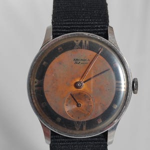 May include: A vintage Arcadia wristwatch with a black strap and a silver case. The watch face is a copper colour with black Roman numeral hour markers and a small sub-dial. The watch face also has the text "Arcadia Fab Suisse" printed on it.