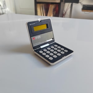 Könnte beinhalten: Ein offener, gebrauchsbereiter Vintage-Taschenrechner Sharp EL-8019. Der Rechner hat ein silbernes und schwarzes Gehäuse mit weißen Tasten und einem gelben Display. Der Rechner steht auf einer weißen Oberfläche.