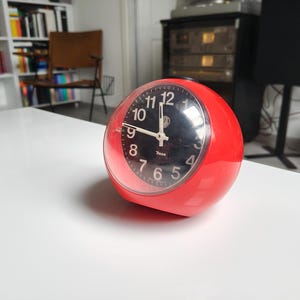 Può includere: Una sveglia analogica sferica rossa con quadrante nero e numeri bianchi è appoggiata su una superficie bianca. L'orologio ha una copertura in plastica trasparente e una base in metallo argentato. L'ora è circa le 9:50.